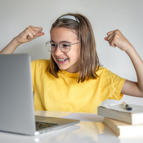 Menina com aparelho nos dentes e camiseta amarela comemorando em frente ao notebook durante uma aula online, com expressão de alegria.