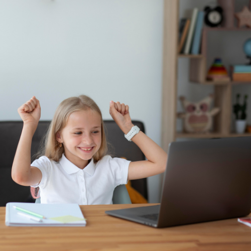 Menina sorrindo e levantando os braços em comemoração durante uma aula online, sentada em frente ao notebook com cadernos sobre a mesa.