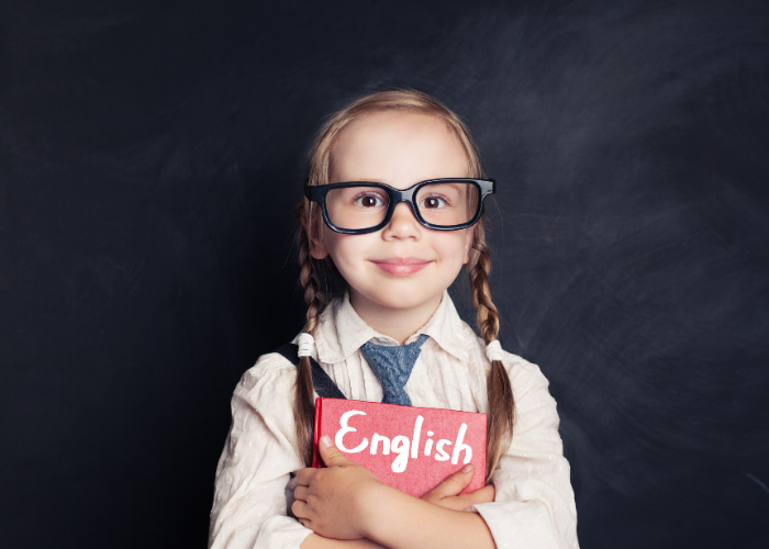 Menina sorridente com óculos grandes segurando um livro vermelho escrito “English”, em frente a um fundo escuro.