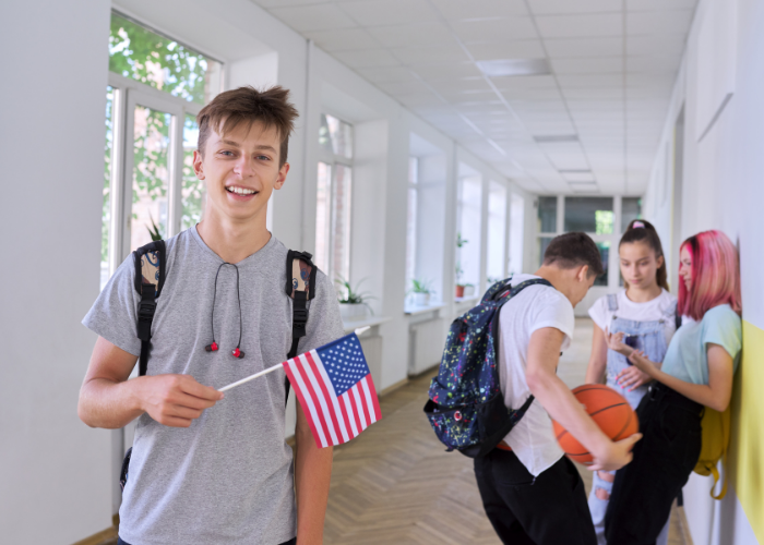 Adolescente segurando uma bandeira dos Estados Unidos em um corredor escolar, representando interesse por intercâmbio e aprendizado de inglês.