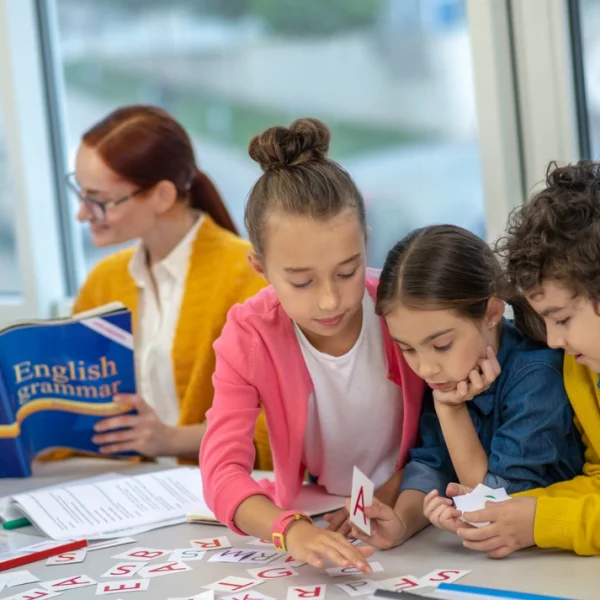 Grupo de crianças trabalhando em equipe com letras de alfabeto na mesa, enquanto uma professora observa ao fundo com um livro de gramática.